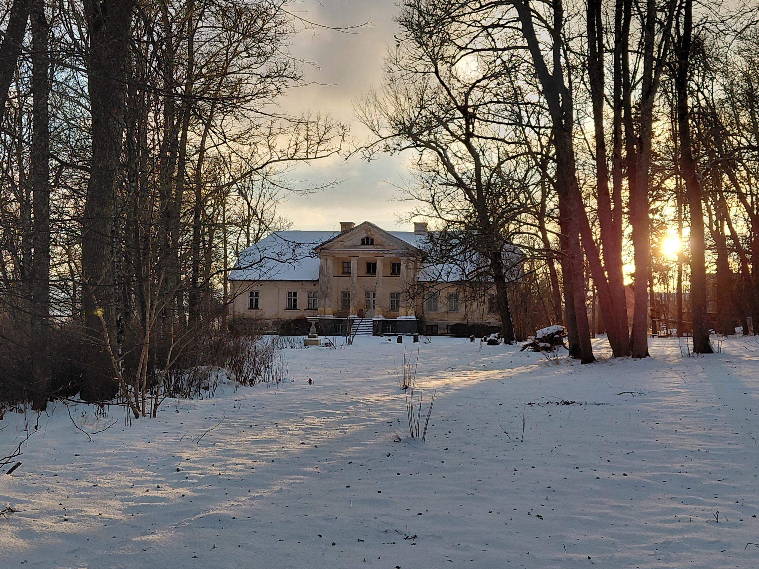 Padures muižas galvenā ēka ziemā, sniegots pagalms un kaili koki saulrieta gaismā.